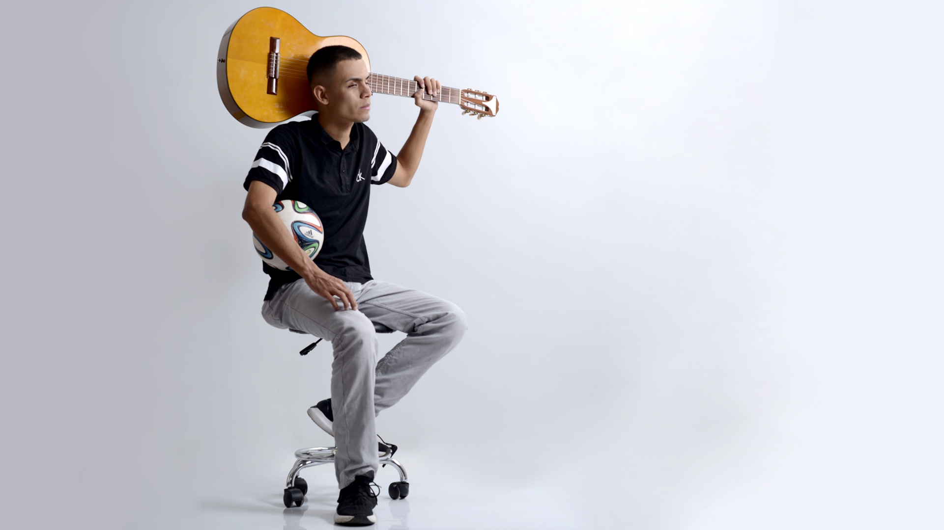 Jose Quiceno sostiene un balón bajo su brazo derecho y su guitarra detrás de su cabeza tomada por el mástil con su mano izquierda Jose Quiceno sostiene un balón bajo su brazo derecho y su guitarra detrás de su cabeza tomada por el mástil con su mano izquierda